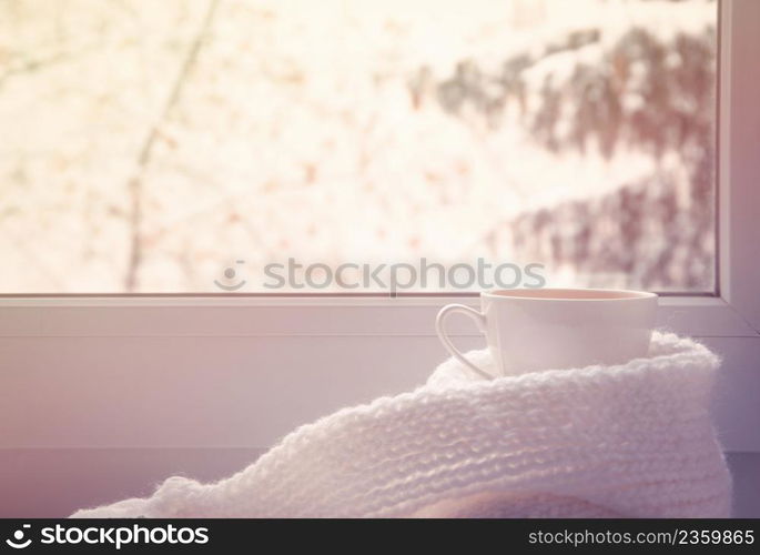 Mug of hot tea and warm woolen knitting on windowsill against snow landscape from outside.