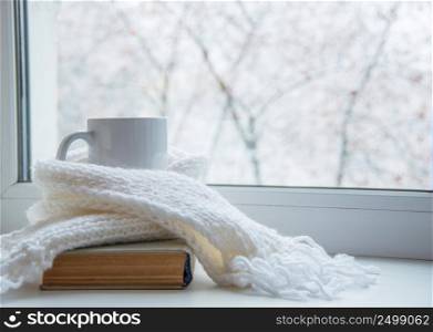 Mug of hot tea and warm woolen knitting on windowsill against snow landscape from outside.