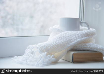 Mug of hot tea and warm woolen knitting on windowsill against snow landscape from outside.