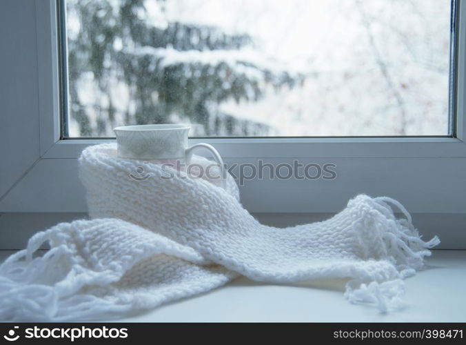 Mug of hot tea and warm woolen knitting on windowsill against snow landscape from outside.