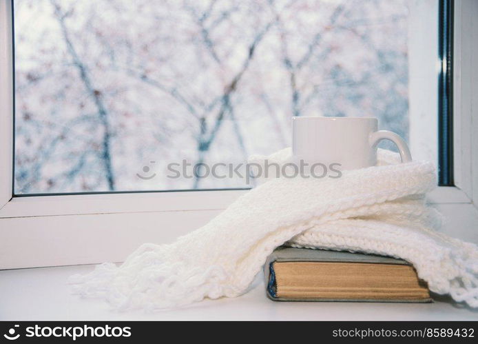 Mug of hot tea and warm woolen knitting on windowsill against snow landscape from outside.