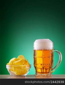 Mug of light beer with foam and chips in a plate on a green background. Mug of light beer with foam and chips