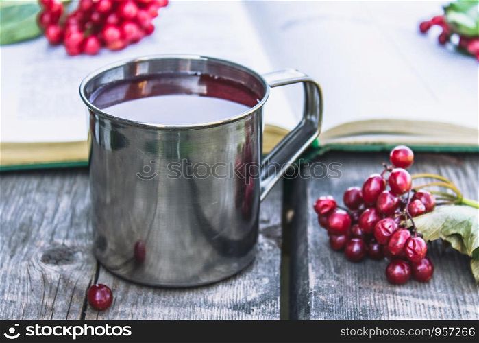 Mug or cup of hot viburnum tea on a wooden table next to an open book and red berries of viburnum. Source of natural vitamins. Used in folk medicine. Close-up.. Mug or cup of hot viburnum tea on a wooden table next to an open book and red berries of viburnum. Source of natural vitamins. Used in folk medicine.