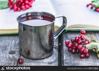 Mug or cup of hot viburnum tea on a wooden table next to an open book and red berries of viburnum. Source of natural vitamins. Used in folk medicine. Close-up.. Mug or cup of hot viburnum tea on a wooden table next to an open book and red berries of viburnum. Source of natural vitamins. Used in folk medicine.