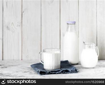 Mug with milk, bottle of milk and jug of milk on gray table, white wooden background with copy space. Breakfast concept, healthy food, reusable glassware. Mug with milk, bottle of milk and jug of milk on a gray table
