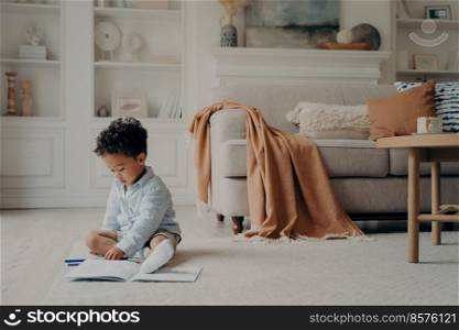 Mulatto curly haired cute boy calmly sitting on warm floor playing with pencils in stylishly furnished living room with sofa and coffee table beside him, creative child activity indoor concept. African cute boy calmly sitting on warm floor and playing with colored felt tip pens