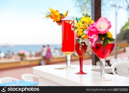 Multi-colored cocktails in high stemware, decorated with flowers and fruit at the bar counter on the beach background. Shallow depth of field