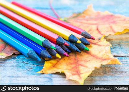 Multi-colored pencils lie on a yellow autumn leaf on a background of old blue boards. School concept. Multi-colored pencils lie on a yellow autumn leaf on a background of old blue boards.