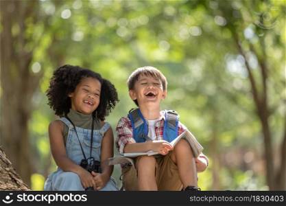 Multi-ethnic children in casual clothing sitting on tree roots, laughing merrily during hiking.Exploring nature and camping summer concept.