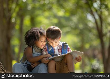 Multi-ethnic children in casual clothing sitting on tree roots, looking at the map with interest.Exploring nature and camping summer concept.