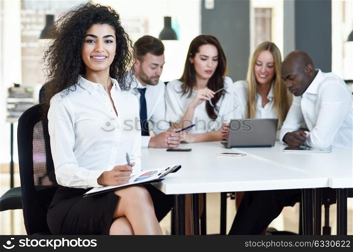 Multi-ethnic group of five businesspeople meeting in a modern office. Muslim businesswoman leader, wearing white shirt and black skirt, looking at camera.