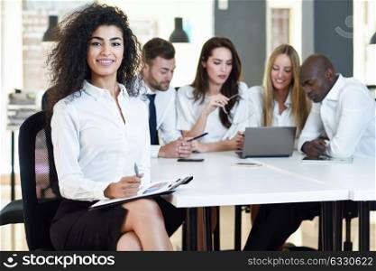 Multi-ethnic group of five businesspeople meeting in a modern office. Muslim businesswoman leader, wearing white shirt and black skirt, looking at camera.