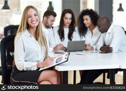 Multi-ethnic group of five businesspeople meeting in a modern office. Caucasian blonde businesswoman leader, wearing white shirt and black skirt, looking at camera.