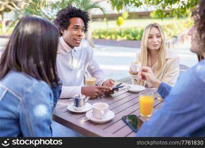 Multi-ethnic group of friends having a drink together in an outdoor bar. One of the men shows something on his smartphone to his friends.. Multi-ethnic group of friends having a drink together in an outdoor bar.