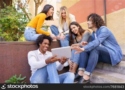 Multi-ethnic group of young people looking at a digital tablet outdoors in urban background. Group of men and woman sitting together on steps.. Multi-ethnic group of young people looking at a digital tablet outdoors in urban background.