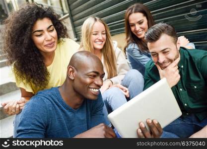 Multi-ethnic group of young people looking at a tablet computer outdoors in urban background. Group of men and woman sitting together on steps.