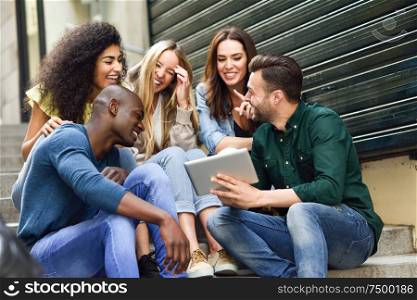 Multi-ethnic group of young people looking at a tablet computer outdoors in urban background. Group of men and woman sitting together on steps.. Multi-ethnic group of young people looking at a tablet computer