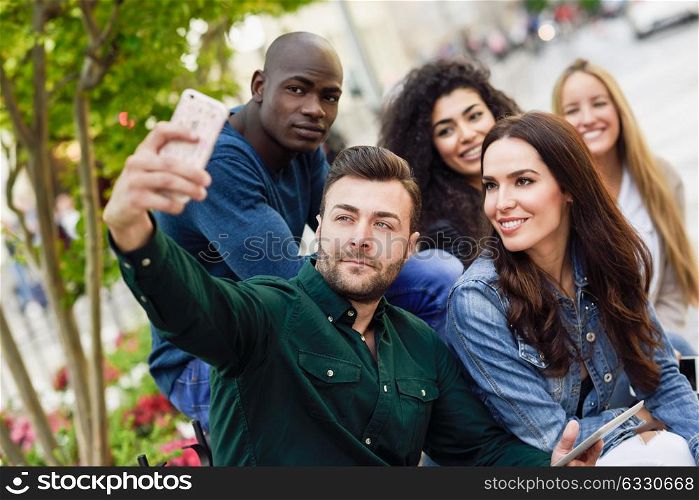 Multi-ethnic group of young people taking selfie photograph together outdoors. Beautiful funny women and men wearing casual clothes in urban background.