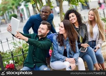 Multi-ethnic group of young people taking selfie photograph together outdoors. Beautiful funny women and men wearing casual clothes in urban background.. Multi-ethnic young people taking selfie together in urban background
