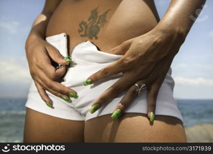 Multi ethnic young adult woman on beach unzipping shorts.