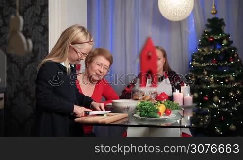 Multi generation family serving Christmas table together. Grandmother teaching her granddaughter how to cut vegetables. Girl cutting cucumber on chopping board and giving her lovely mother a slice to taste over Christmas decorations background.