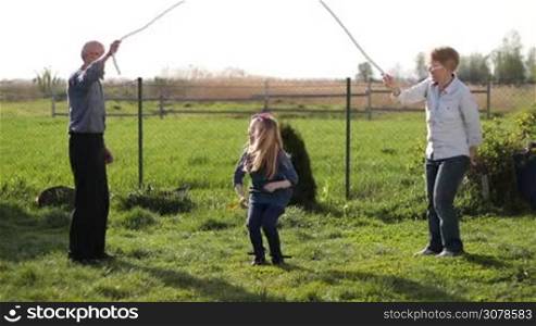 Multi generation family spending leisure in countryside in spring. Adorable blonde girl in eyeglasses jumping over the rope while her grandparents rotating rope outside on green lawn. Little girl having fun outdoors with skipping rope. Slow motion.