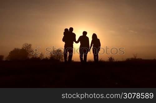 Multi generation family with two children walking into sunset in the meadow. Grandfather carrying toddler boy and grandmother holding granddaughter by hand going into the rays of setting sun. Slow motion. Steadicam stabilized shot.