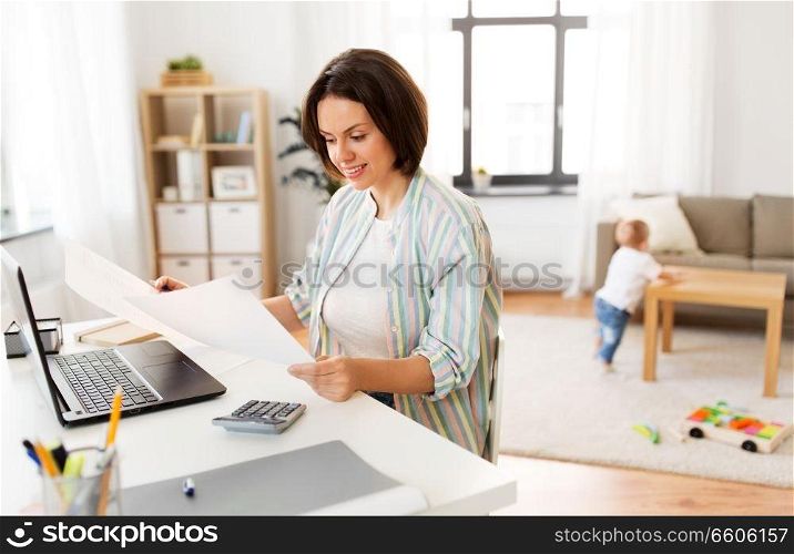 multi-tasking, freelance and motherhood concept - working mother with papers at laptop computer and baby boy at home office. mother working with papers and baby boy at home