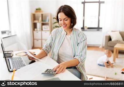 multi-tasking, freelance and motherhood concept - working mother with papers counting on calculator and baby boy playing at home office. working mother counting on calculator and baby