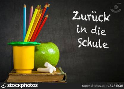 Multicolor pencils, chalks and green apple on old book against blackboard with text &acute;Back to school!&acute; in the German language. School concept
