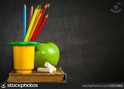 Multicolor pencils, chalks and green apple on old book against blackboard with copy space. School concept