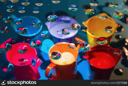 Multicolored abstract background - view through glass with drops of liquid on a blurry colorful watering cans and buckets. Selective focus on foreground.. Multicoloredl abstract background through drops