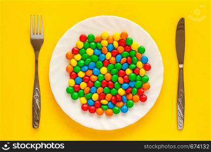 Multicolored candy dragee on a white plate with a knife and fork on a yellow background.