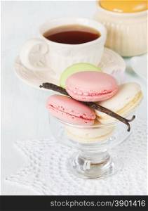 Multicolored macaroons in a glass ice-cream bowl on a round lacy napkin and a cup of tea