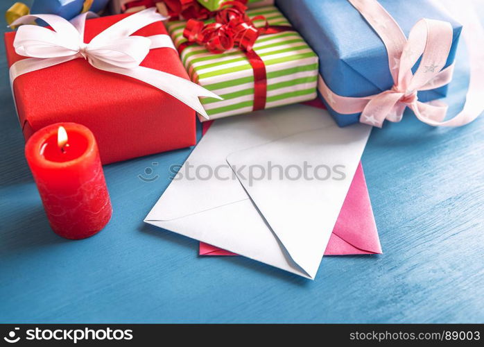 Multicolored presents, wrapped in paper and tied with ribbon and bow, displayed on a wooden table, empty envelopes and a red, lit candle.