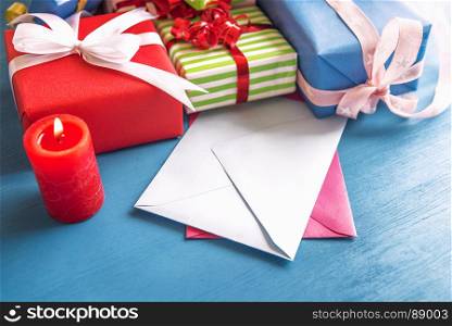 Multicolored presents, wrapped in paper and tied with ribbon and bow, displayed on a wooden table, empty envelopes and a red, lit candle.