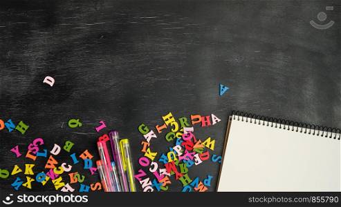multicolored small wooden letters of the English alphabet and paper notebook on a black chalk board, copy space, back to school