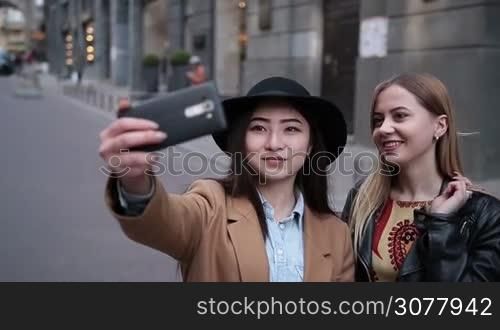 Multiethnic beautiful girlfriends having fun in city taking selfie on the street. Two young pretty females taking selfie with smartphone over store window background. Smiling women making self portrait with mobile phone.