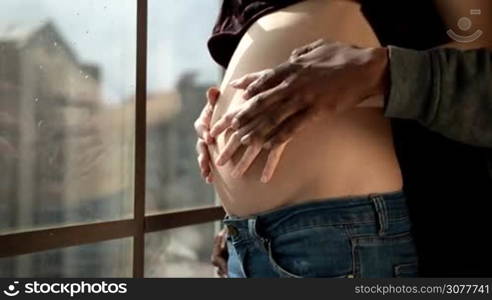 Multiethnic couple expecting a baby. Couple in love touching pregnant woman&acute;s belly with hands against wide window background. Closeup hands of african american father and caucasian mother caressing pregnant belly. Happy family lifestyle.