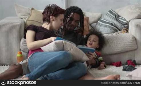 Multiethnic family with curly mixed race son reading fairy tale together while relaxing on carpet near couch at home. African american father with dreadlocks cuddling his little toddler boy while caucasian mother reading book. Slow motion.