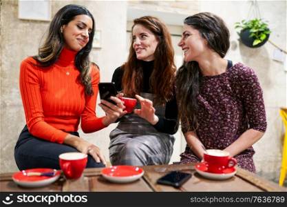 Multiethnic group of three happy female friends looking to a smartphone in a cafe bar.. Group of three happy friends drinking coffee in a cafe bar.