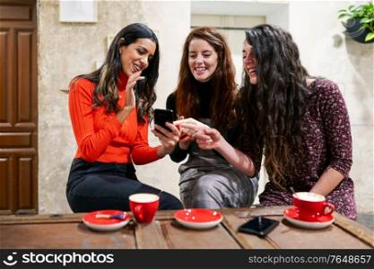 Multiethnic group of three happy female friends looking to a smartphone in a cafe bar.. Group of three happy friends drinking coffee in a cafe bar.
