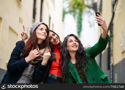 Multiethnic group of three happy woman taking a selfie photo with a smartphone in urban background. Group of three happy woman walking together outdoors