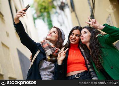 Multiethnic group of three happy woman taking a selfie photo with a smartphone in urban background. Group of three happy woman walking together outdoors