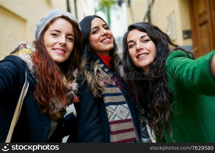 Multiethnic group of three happy woman taking a selfie photo with a smartphone in urban background. Group of three happy woman walking together outdoors