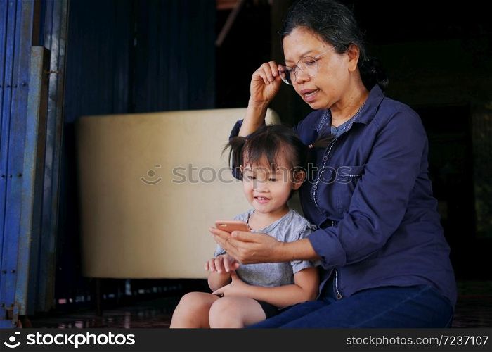 Multigeneration togetherness in Asian family, little girl watching smart phone and listening story tale with grandmother in countryside home.
