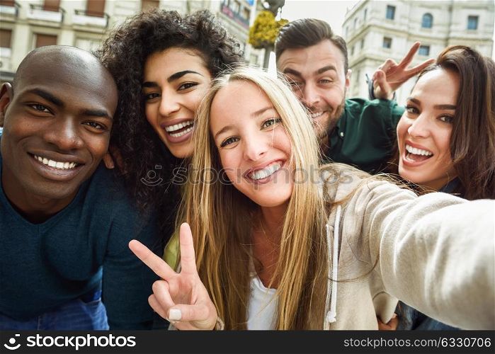 Multiracial group of friends taking selfie in a urban street with a blonde woman in foreground. Three young women and two men wearing casual clothes.