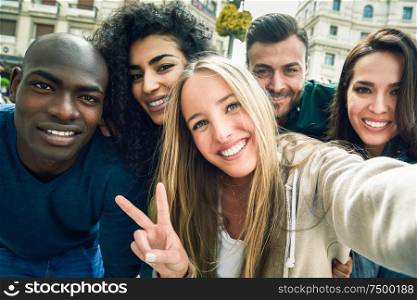 Multiracial group of friends taking selfie in a urban street with a blonde woman in foreground. Three young women and two men wearing casual clothes.. Multiracial group of young people taking selfie
