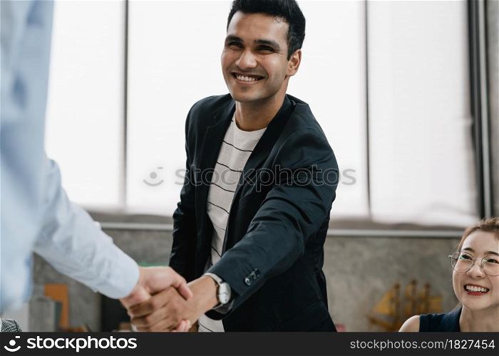 Multiracial group of young creative people in smart casual wear discussing business shaking hands together and smiling while sitting in modern office. Partner cooperation, coworker teamwork concept.