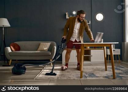 Multitasking businessman in suit and home clothes vacuuming and working online using laptop. Glad multitasking businessman vacuuming and working online